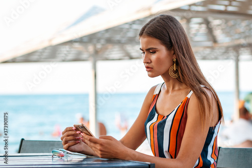 A woman with a concerned expression uses her smartphone. Summer cafe on the beach. Concept of business and online technologies