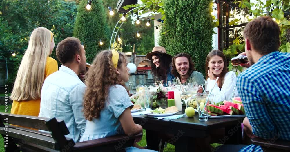 Many mixed race young people sitting at table and talking in garden. African American and Caucasian beautiful girls smiling outdoor. Joyful Asian girl and handsome males chatting. Celebration concept