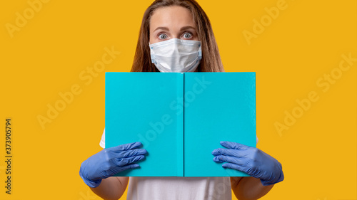 School reopen. Quarantine class. Scared female student in protecting face mask gloves holding open blue book isolated on bright yellow copy space. COVID-19 outbreak. Hygiene measures. Safe education.