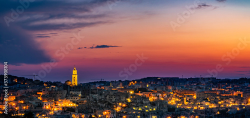 The spectacular fiery sunset and the panorama of the Sassi di Matera in Basilicata, Italy. The top view of the city from the park of the Rupestrian Churches. The lights come on as night falls.