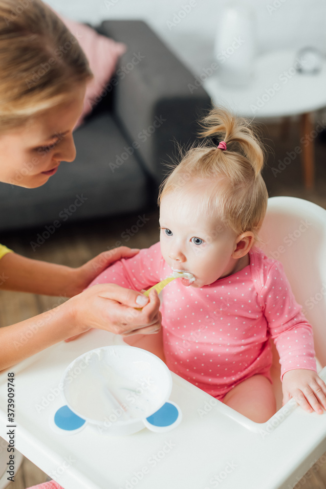 Selective focus of mother feeding baby daughter on highchair at home