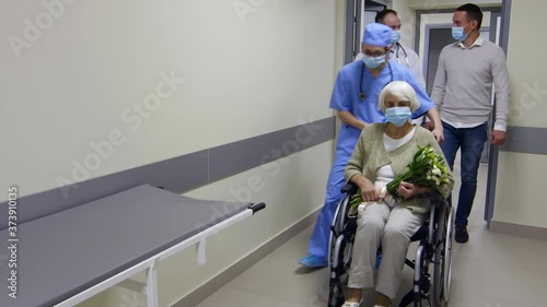 Senior woman in protective face mask holding flowers and riding wheelchair with help of healthcare worker while leaving hospital; her son shaking hands and speaking with doctor
