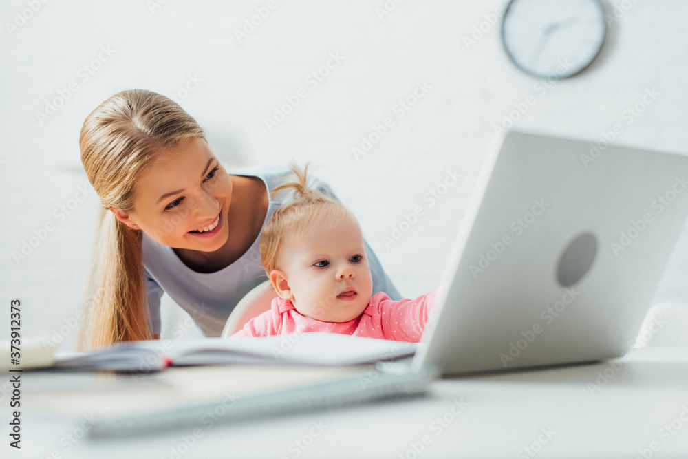 Selective focus of mother standing near kid looking at laptop and stationery on table Selective focus of mother standing near kid looking at laptop and stationery on table