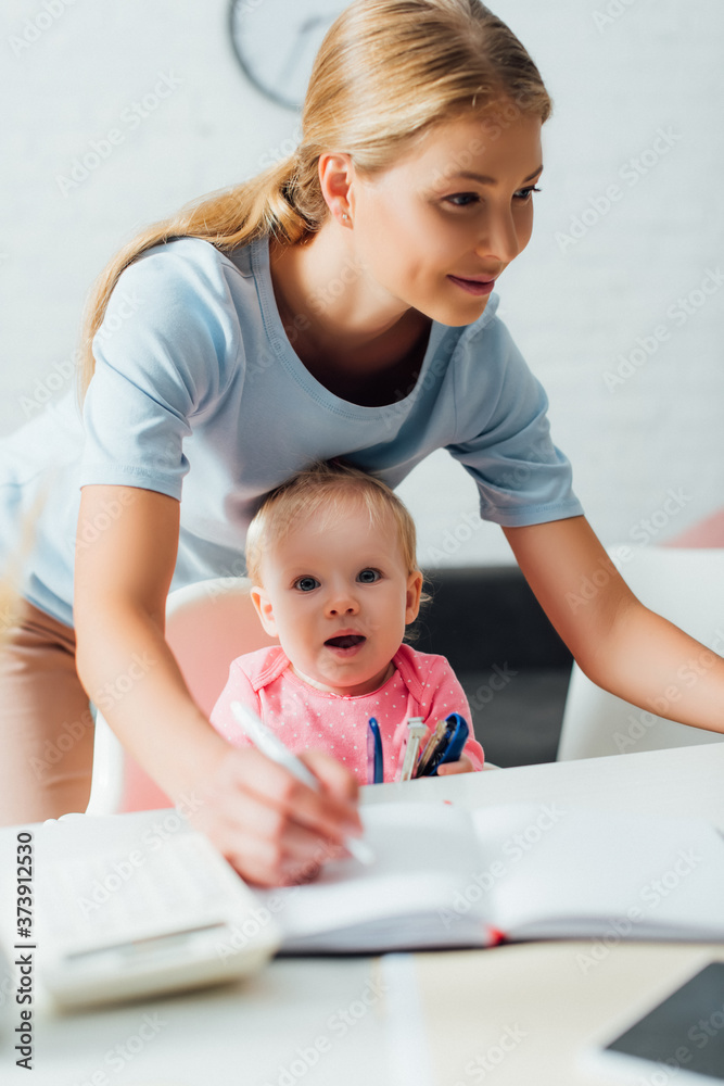 Selective focus of child holding stapler while mother writing on notebook at home Selective focus of child holding stapler while mother writing on notebook at home