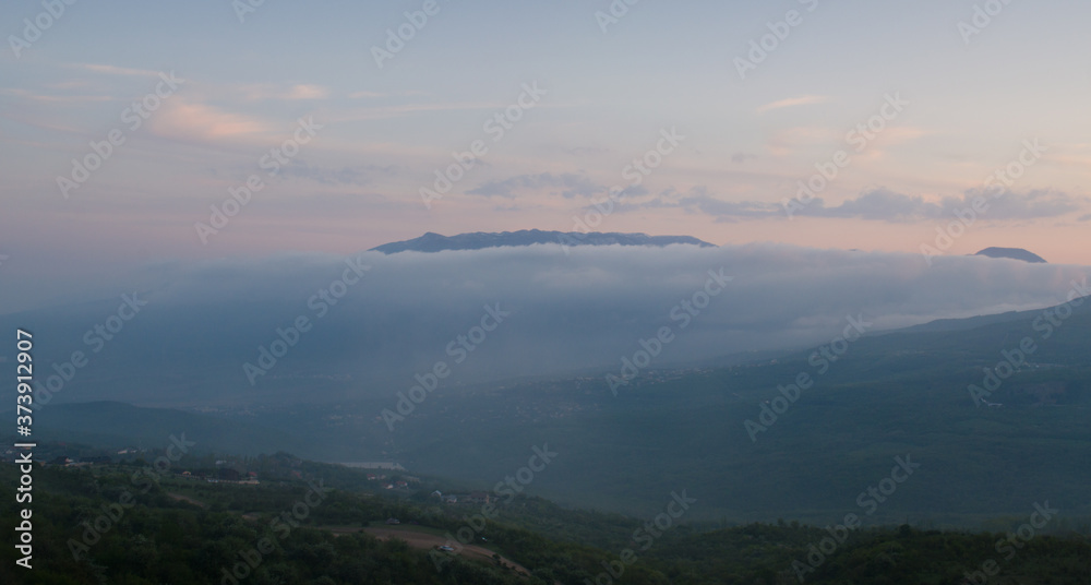 Fog covers the mountains in the evening twilight, Crimea. The lonely road climbs the hill, the houses are lost in the green. Tranquil picturesque evening valley, Scenery of nature of Southern Crimea.