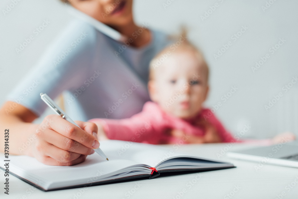 Selective focus of woman writing on notebook and talking on smartphone near child at home Selective focus of woman writing on notebook and talking on smartphone near child at home