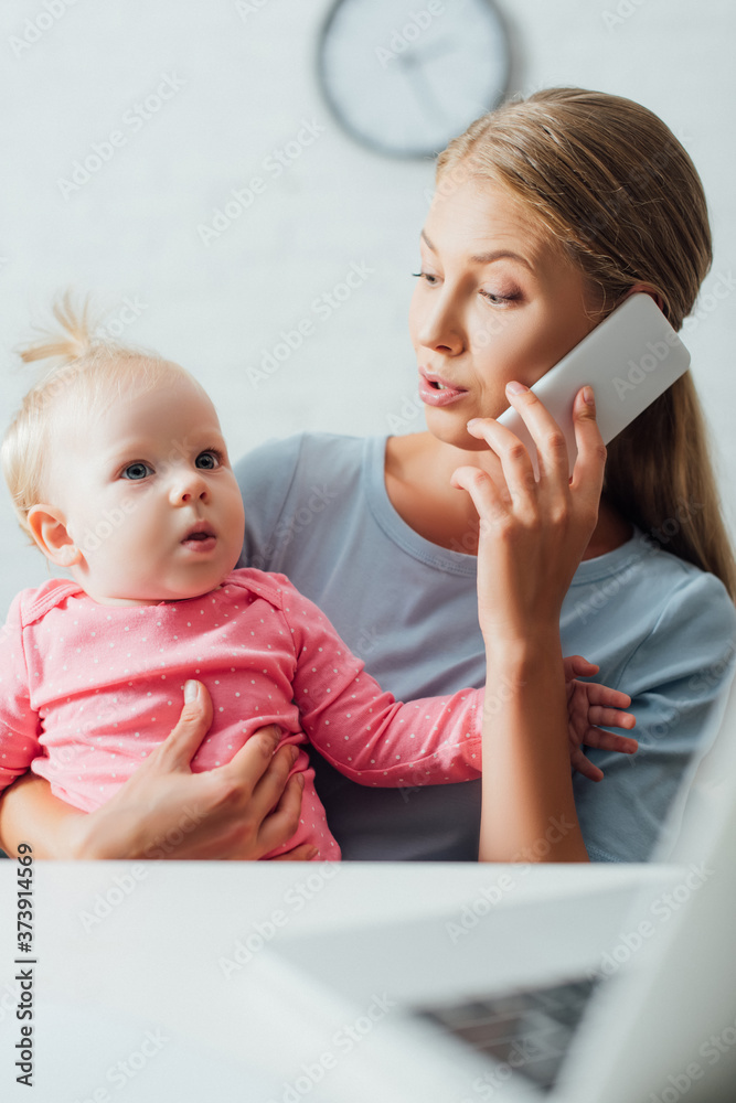 Selective focus of mother holding baby girl and talking on smartphone while working at home Selective focus of mother holding baby girl and talking on smartphone while working at home