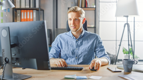 Creative Entrepreneur Sitting at His Desk Works on Desktop Computer in the Stylish Office. Handsome Young Businessman Uses Computer, Does Outsourcing Job, Designs New Apps and Develops Software