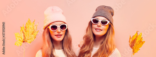 Two fashionable hipster woman sisters in Trendy autumn fall outfit, stylish hair, makeup. Friends in jumper, fashion jeans having fun. Beautiful girl in autumnal beanie hat with maple leaf