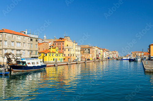 Narrow water canal with boats and fishing ships moored near embankment and colorful buildings in Chioggia town historical centre, blue sky background in summer day, Veneto Region, Northern Italy