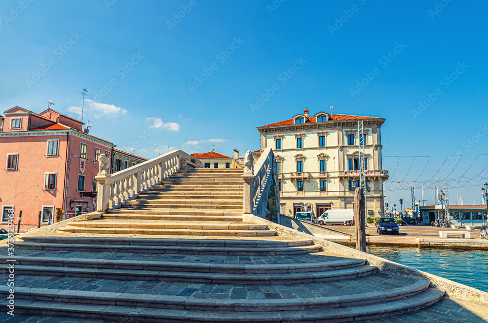 Stone bridge Ponte di Vigo with stairs across Vena water canal and old buildings in historical centre of Chioggia town, blue sky background in summer day, Veneto Region, Northern Italy