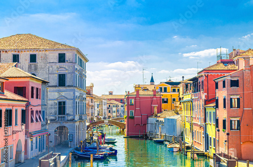 Chioggia cityscape with narrow water canal Vena with moored multicolored boats between old colorful buildings and brick bridge  blue sky background in summer day  Veneto Region  Northern Italy