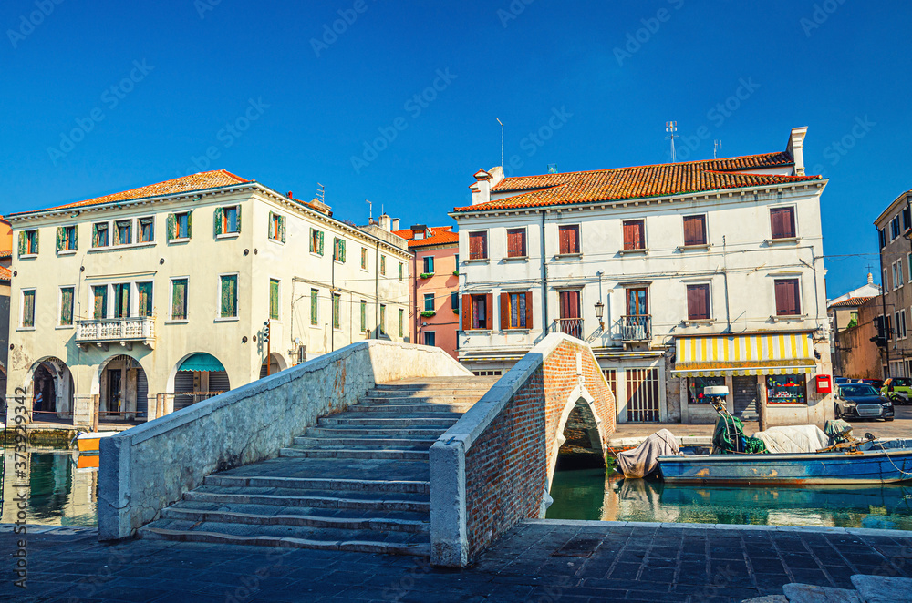 Stone brick bridge Ponte Pescheria across Vena water canal and old buildings in historical centre of Chioggia town, blue sky background in summer day, Veneto Region, Northern Italy