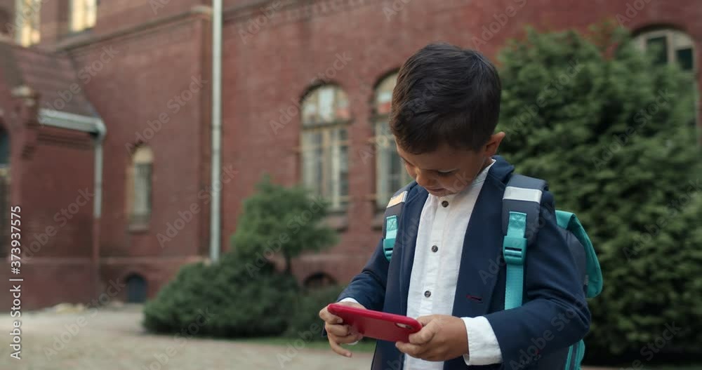 Kid making face while playing online game on smartphone at street. Boy in uniform with backpack touching phone screen while standing near school. Concept of gaming and children. Kid making face while playing online game on smartphone at street. Boy in uniform with backpack touching phone screen while standing near school. Concept of gaming and children.