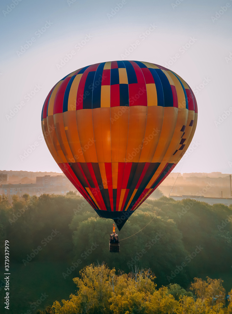 Fototapeta premium Colorful hot air balloon takes off from green park in small european city at summer sunrise