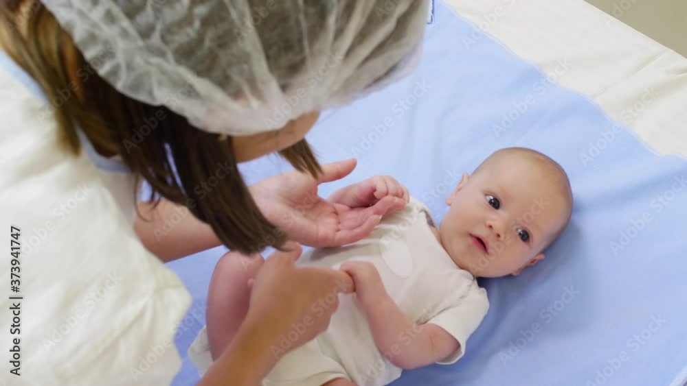 High angle view of woman in disposable hat holding hands of cute newborn baby and playing with him on medical couch High angle view of woman in disposable hat holding hands of cute newborn baby and playing with him on medical couch