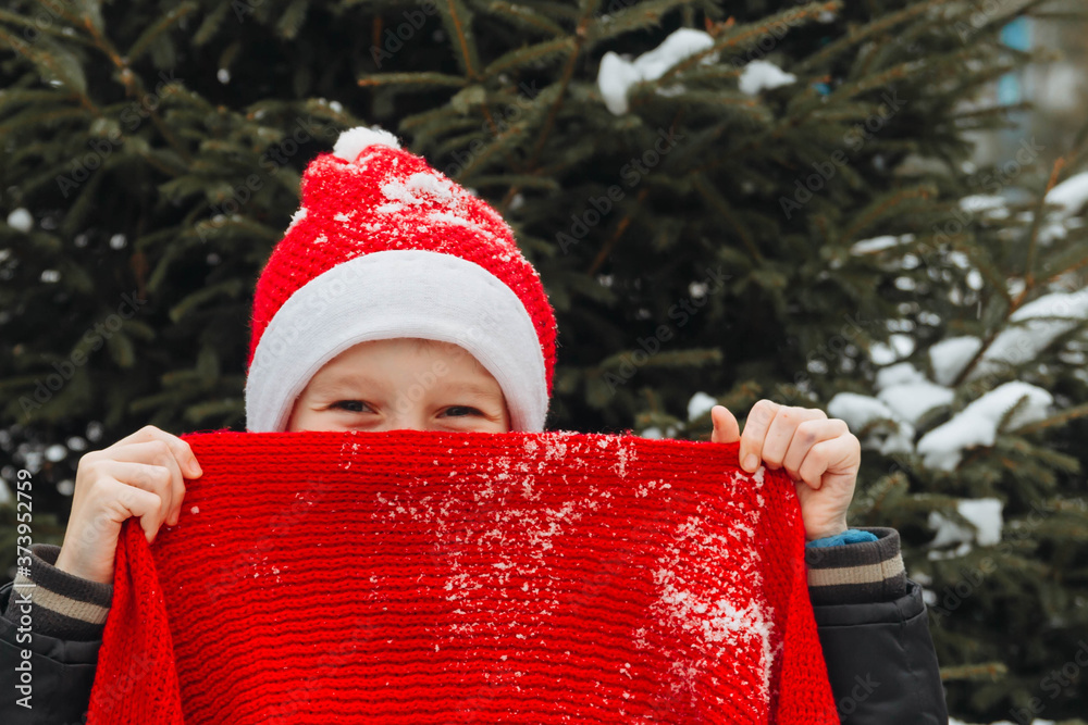 Obraz premium A teenage European boy in a red Santa suit hides behind a bright festive knitted scarf. Happy face of a child close-up. Background for Christmas.