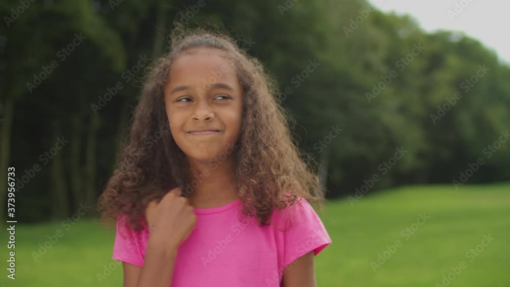 Outdoor portrait of shy adorable elementary age african american girl twisting her curly hair on finger, thinking about escapade, looking with innocent modest smile while standing in summer nature.