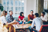 Joyful people dressed in casual wear playing cards game at table with drinks during friendly meeting in comfortable flat, happy multicultural hipsters having fun spending weekend for poker gambling