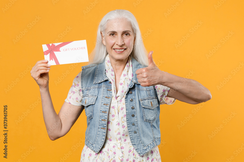 Smiling elderly gray-haired female woman wearing casual dress denim waistcoat posing hold gift certificate showing thumb up looking camera isolated on yellow color wall background studio portrait. Smiling elderly gray-haired female woman wearing casual dress denim waistcoat posing hold gift certificate showing thumb up looking camera isolated on yellow color wall background studio portrait.