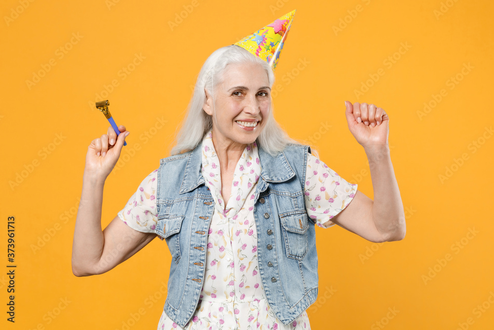 Obraz premium Smiling elderly gray-haired female woman 60s 70s wearing casual dress denim waistcoat birthday hat celebrating hold pipe dancing clenching fists isolated on yellow color background studio portrait.
