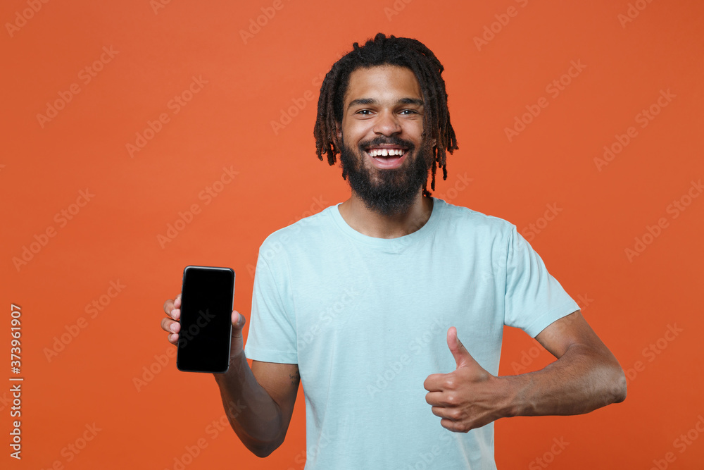 Funny young african american man guy in blue casual t-shirt isolated on orange background. People lifestyle concept. Mock up copy space. Hold mobile phone with blank empty screen, showing thumb up. - obrazy, fototapety, plakaty 