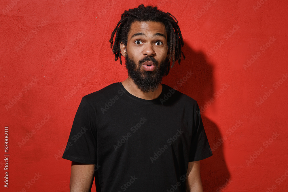 Naklejka premium Shocked amazed surprised young african american man guy with dreadlocks 20s wearing black casual t-shirt posing saying wow looking camera isolated on bright red color wall background studio portrait.