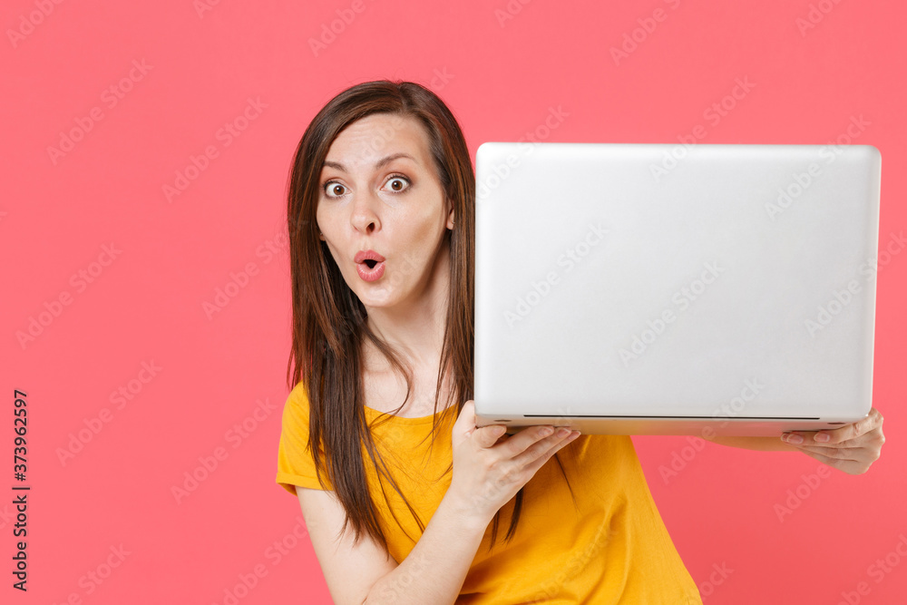 Shocked amazed young brunette woman 20s wearing yellow casual t-shirt posing standing and working on laptop pc computer say wow looking camera isolated on pink color wall background studio portrait. Shocked amazed young brunette woman 20s wearing yellow casual t-shirt posing standing and working on laptop pc computer say wow looking camera isolated on pink color wall background studio portrait.