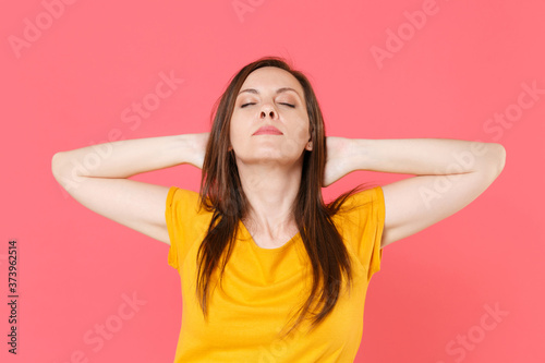 Relaxed beautiful attractive young brunette woman 20s wearing yellow casual t-shirt posing sleeping with hands behind head keeping eyes closed isolated on pink color wall background studio portrait.