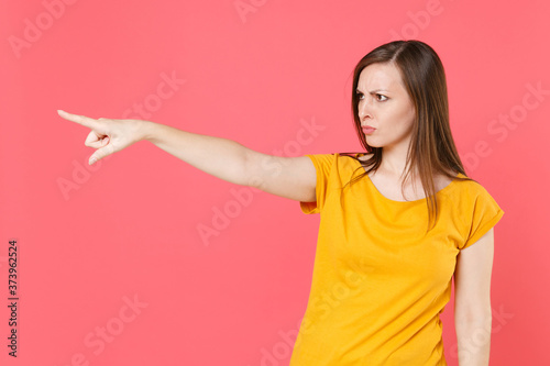 Displeased dissatisfied disconcerted strict young brunette woman 20s wearing yellow casual t-shirt posing standing pointing index finger aside isolated on pink color wall background studio portrait.