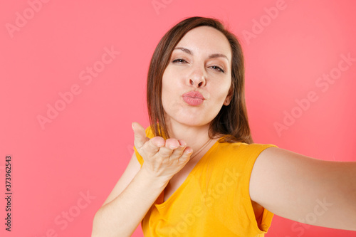 Close up of pretty charming young brunette woman 20s wearing yellow casual t-shirt posing doing selfie shot on mobile phone blowing sending air kiss isolated on pink color background studio portrait.