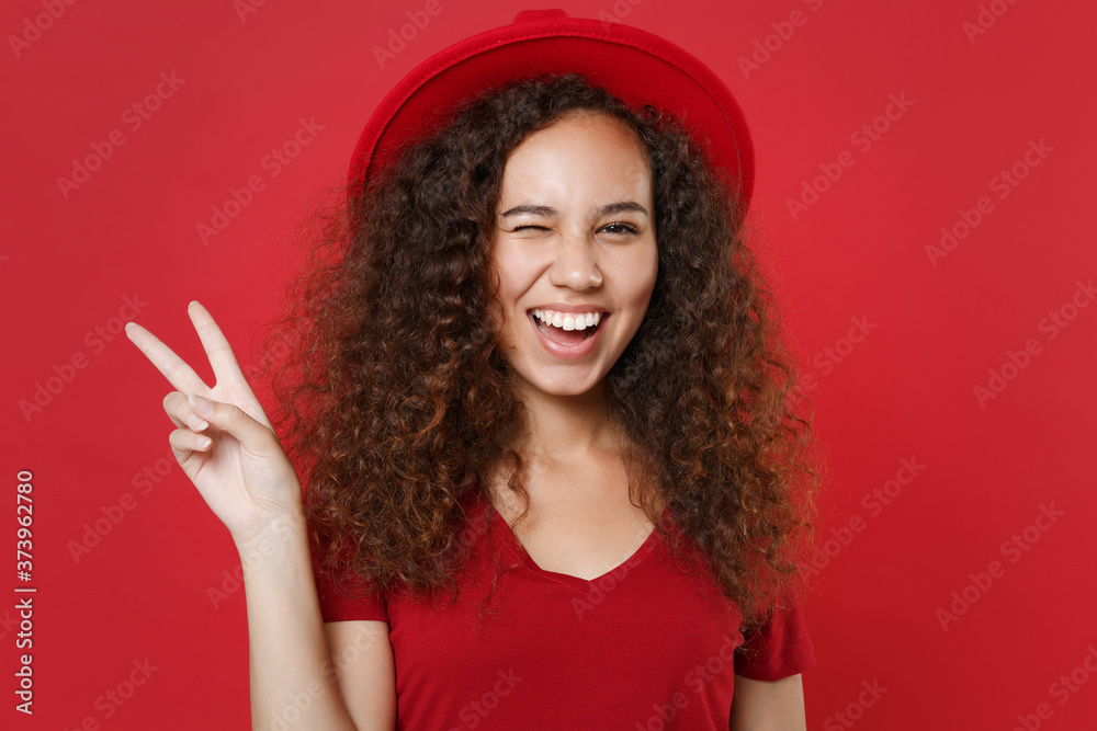 Blinking young african american woman girl in casual t-shirt hat posing isolated on bright red background studio portrait. People emotions lifestyle concept. Mock up copy space. Showing victory sign. - obrazy, fototapety, plakaty 