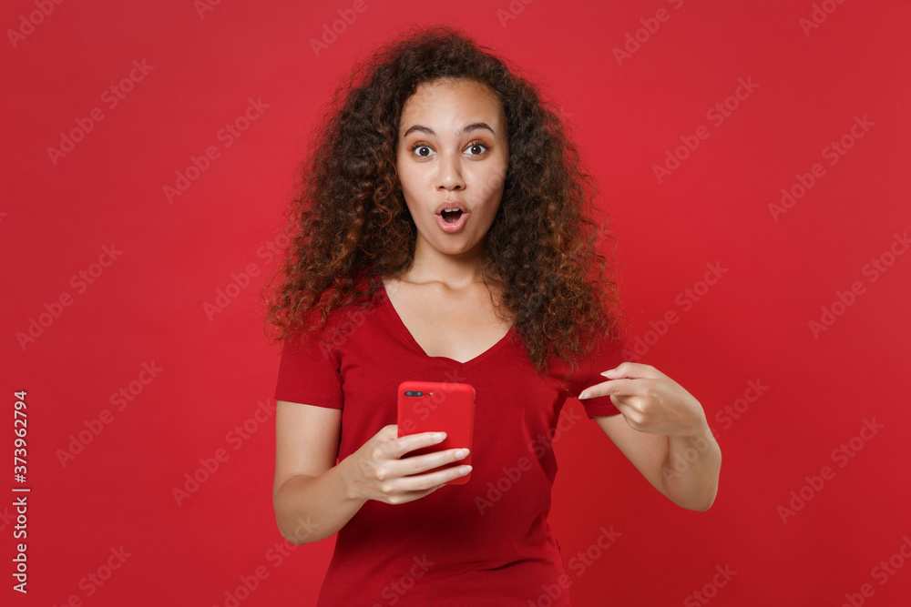Shocked young african american girl in casual t-shirt posing isolated on red wall background studio portrait. People lifestyle concept. Mock up copy space. Pointing index finger on mobile cell phone. Shocked young african american girl in casual t-shirt posing isolated on red wall background studio portrait. People lifestyle concept. Mock up copy space. Pointing index finger on mobile cell phone.