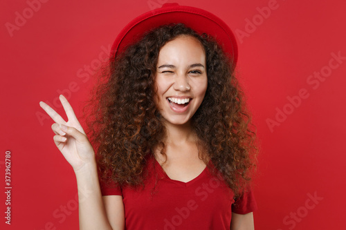 Blinking young african american woman girl in casual t-shirt hat posing isolated on bright red background studio portrait. People emotions lifestyle concept. Mock up copy space. Showing victory sign.