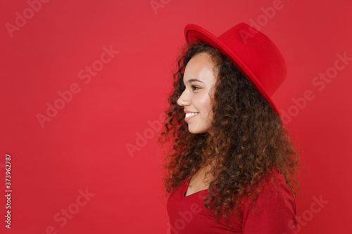 Side view of smiling young african american woman girl in casual t-shirt hat posing isolated on bright red wall background studio portrait. People lifestyle concept. Mock up copy space. Looking aside.