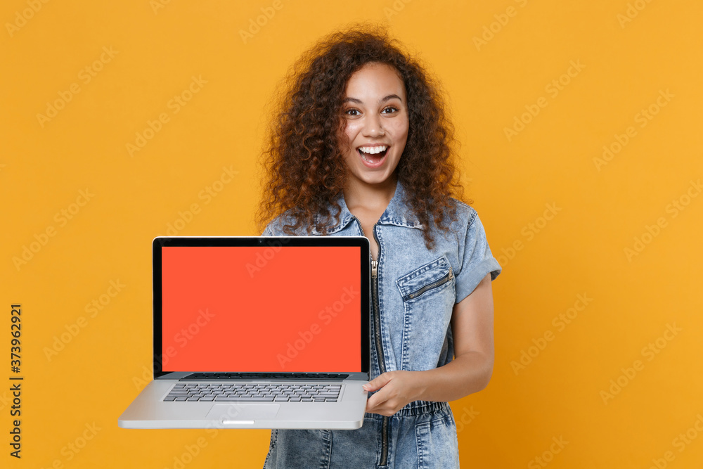 Naklejka premium Surprised young american girl in casual denim clothes isolated on yellow wall background studio portrait. People lifestyle concept. Mock up copy space. Hold laptop pc computer with blank empty screen.