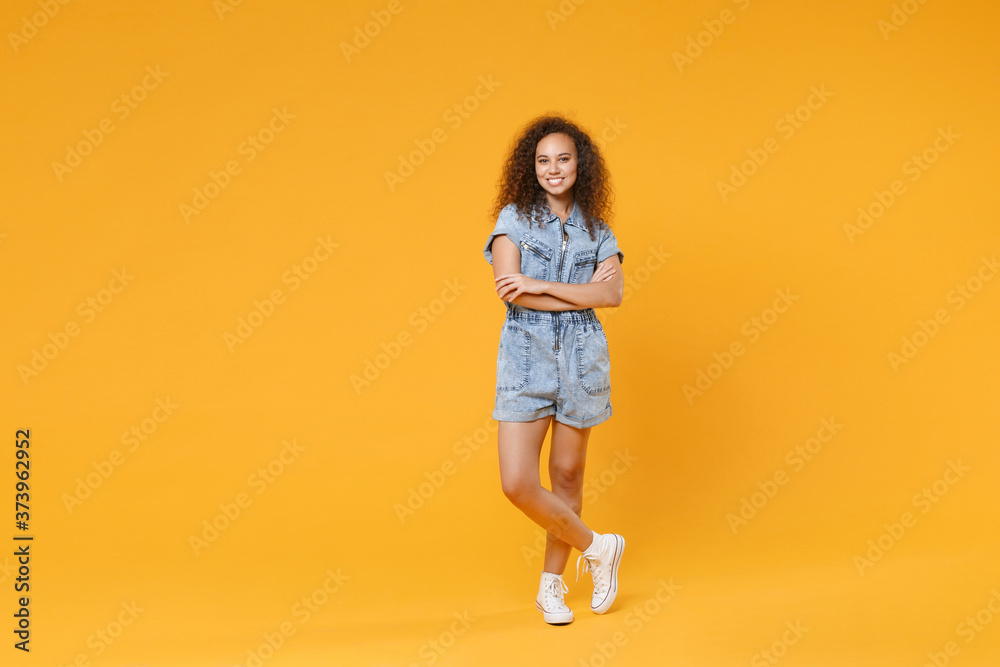 Full length portrait of smiling young african american girl in denim clothes isolated on yellow wall background studio portrait. People lifestyle concept. Mock up copy space. Holding hands crossed. Full length portrait of smiling young african american girl in denim clothes isolated on yellow wall background studio portrait. People lifestyle concept. Mock up copy space. Holding hands crossed.