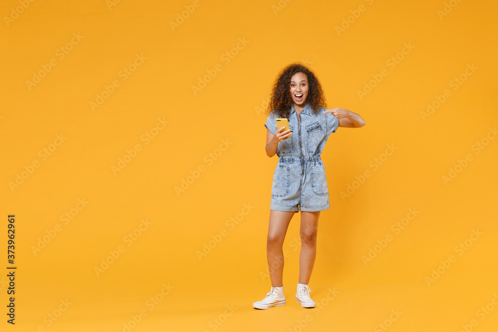 Full length portrait of excited young african american woman girl in denim clothes isolated on yellow wall background in studio. People lifestyle concept. Pointing index finger on mobile cell phone.