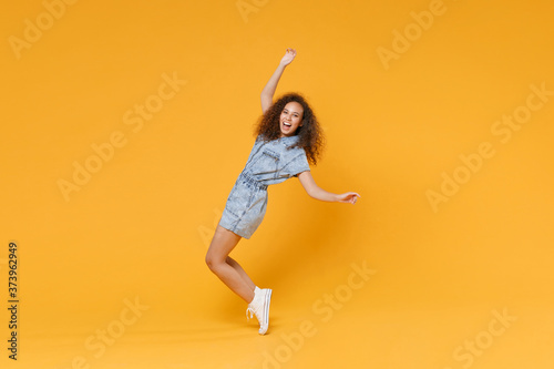 Full length portrait of cheerful young african american woman girl in denim clothes isolated on yellow background. People lifestyle concept. Mock up copy space. Dancing standing on toes, rising hands.