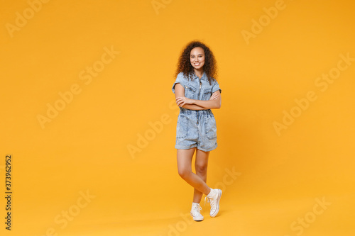 Full length portrait of smiling young african american girl in denim clothes isolated on yellow wall background studio portrait. People lifestyle concept. Mock up copy space. Holding hands crossed.