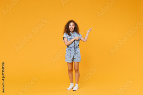Full length portrait of surprised young african american woman girl in denim clothes isolated on yellow background studio portrait. People emotions lifestyle concept. Pointing index fingers aside.