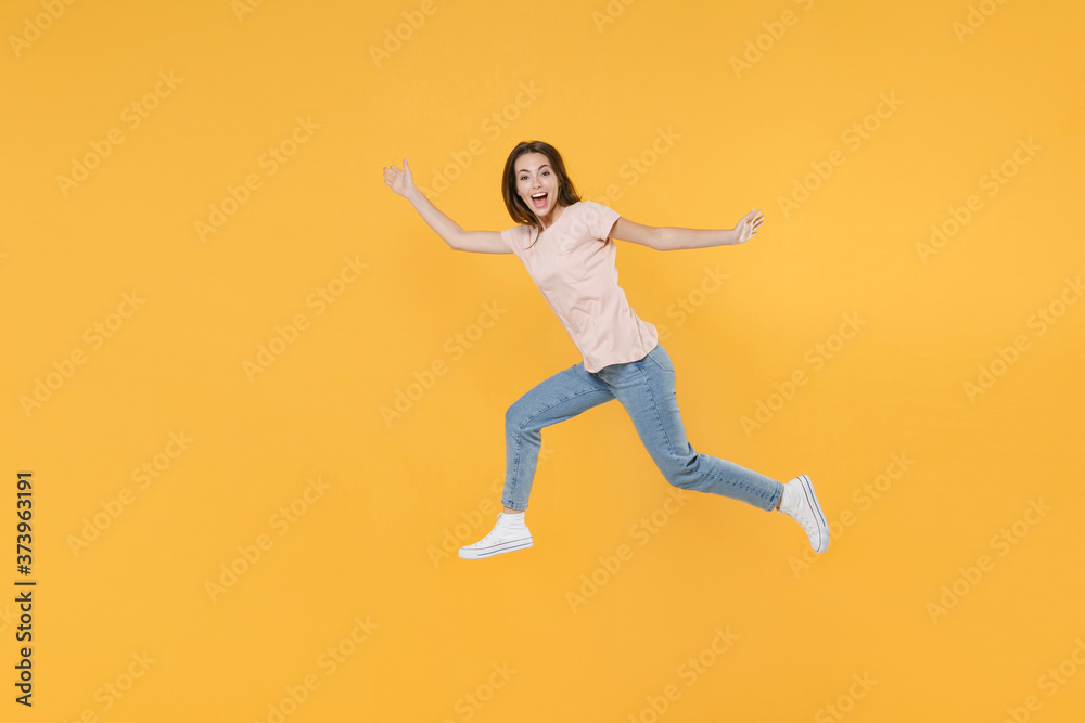 Full length side view portrait of cheerful funny young woman 20s wearing pastel pink casual t-shirt posing jumping spreading hands and legs looking camera isolated on yellow color background studio.