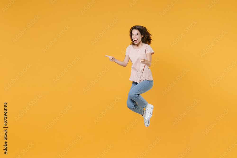 Full length portrait of excited cheerful young brunette woman 20s wearing pastel pink casual t-shirt posing jumping pointing index fingers aside isolated on bright yellow color wall background studio.