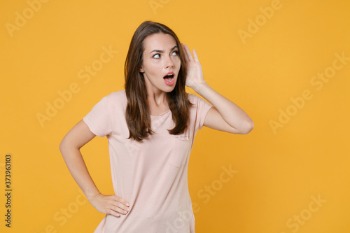 Shocked curious young brunette woman 20s wearing pastel pink casual t-shirt posing try to hear you overhear listening intently looking aside isolated on yellow color wall background studio portrait.