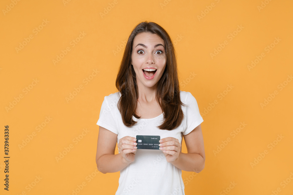 Shocked amazed excited young brunette woman 20s wearing white blank casual t-shirt posing hold credit bank card keeping mouth open looking camera isolated on yellow color background studio portrait. Shocked amazed excited young brunette woman 20s wearing white blank casual t-shirt posing hold credit bank card keeping mouth open looking camera isolated on yellow color background studio portrait.