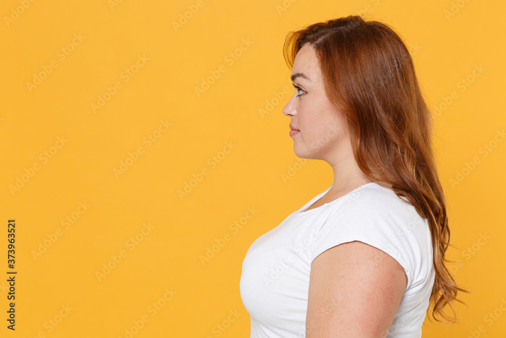 Side view of smiling pretty young redhead plus size body positive female woman girl 20s in white blank design casual t-shirt posing looking aside isolated on yellow color background studio portrait. Side view of smiling pretty young redhead plus size body positive female woman girl 20s in white blank design casual t-shirt posing looking aside isolated on yellow color background studio portrait.