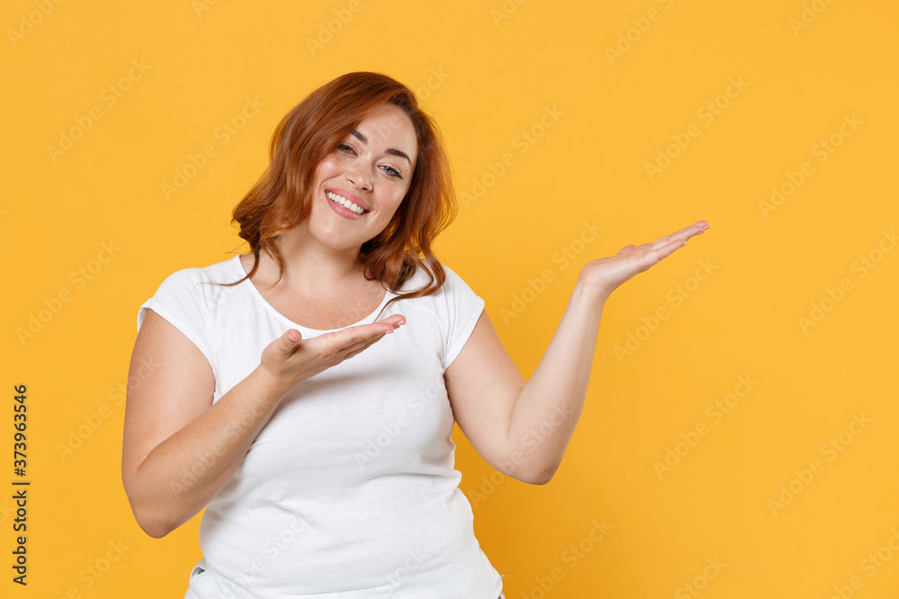 Smiling young redhead plus size body positive female woman girl 20s in white blank casual t-shirt posing pointing hands aside on mock up copy space isolated on yellow color background studio portrait. Smiling young redhead plus size body positive female woman girl 20s in white blank casual t-shirt posing pointing hands aside on mock up copy space isolated on yellow color background studio portrait.