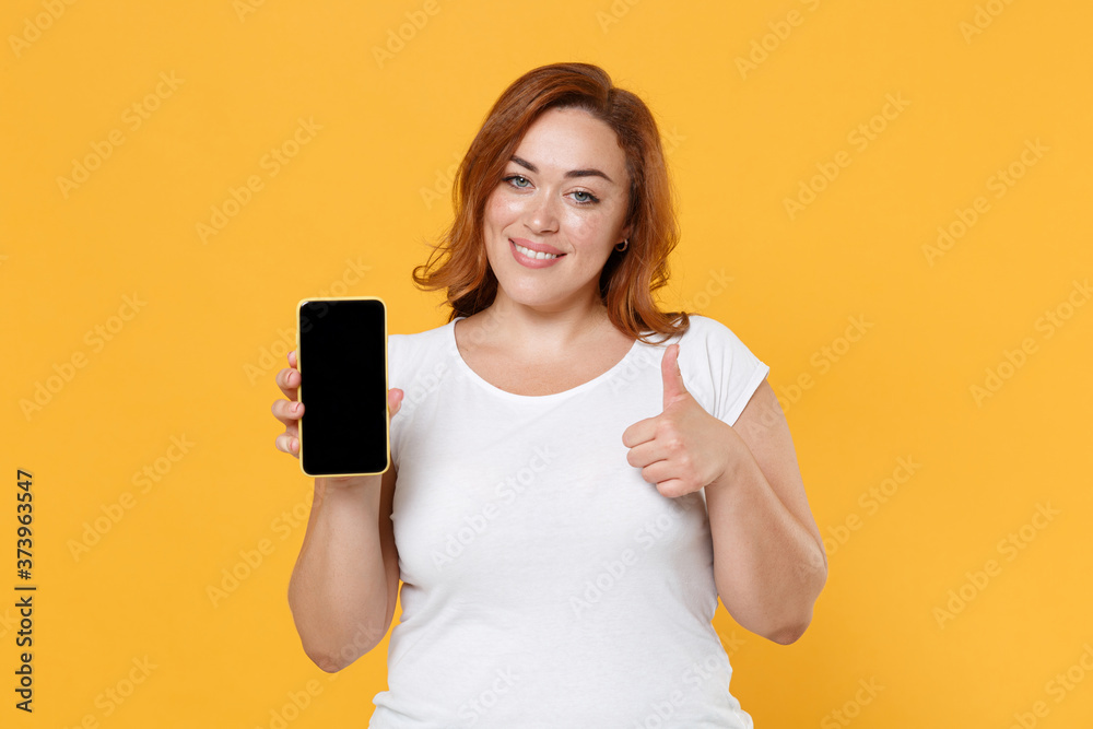 Smiling young redhead plus size body positive female woman in white t-shirt hold mobile phone with blank empty screen mock up copy space showing thumb up isolated on yellow background studio portrait. Smiling young redhead plus size body positive female woman in white t-shirt hold mobile phone with blank empty screen mock up copy space showing thumb up isolated on yellow background studio portrait.