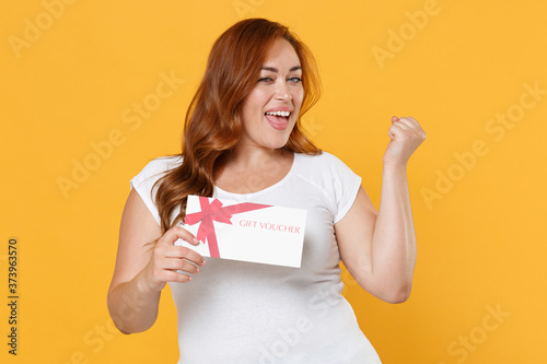 Joyful young redhead plus size body positive female woman girl 20s in white blank casual t-shirt posing hold gift certificate doing winner gesture isolated on yellow color background studio portrait.