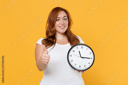 Smiling young redhead plus size body positive female woman girl 20s in white casual t-shirt posing holding clock showing thumb up looking camera isolated on yellow color background studio portrait.